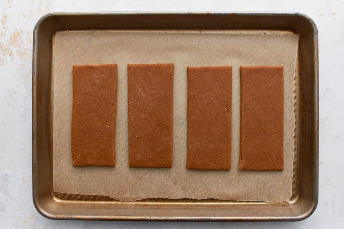 A baking sheet with four large, rectangular pieces of graham cracker dough placed on parchment paper, ready to be scored and baked.