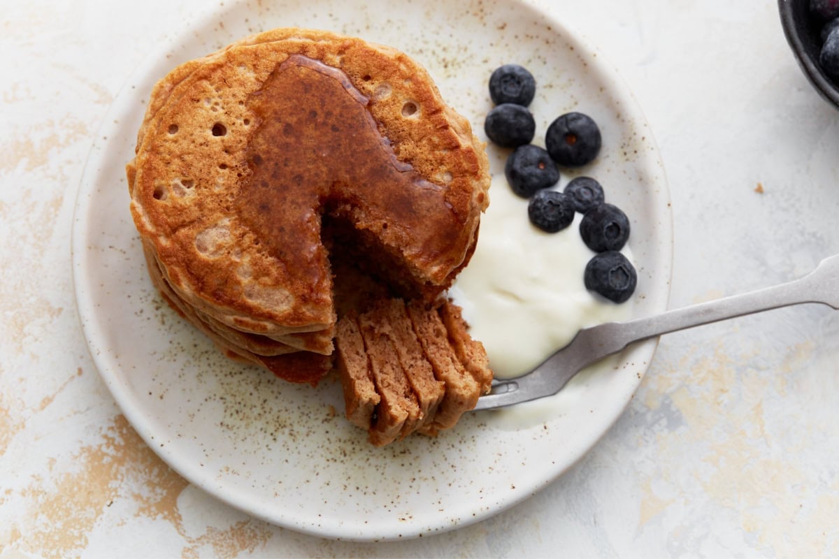 A close-up of a stack of buckwheat pancakes with a cut-out section, revealing their soft, fluffy texture. The plate is garnished with fresh blueberries, yogurt, and a drizzle of maple syrup.