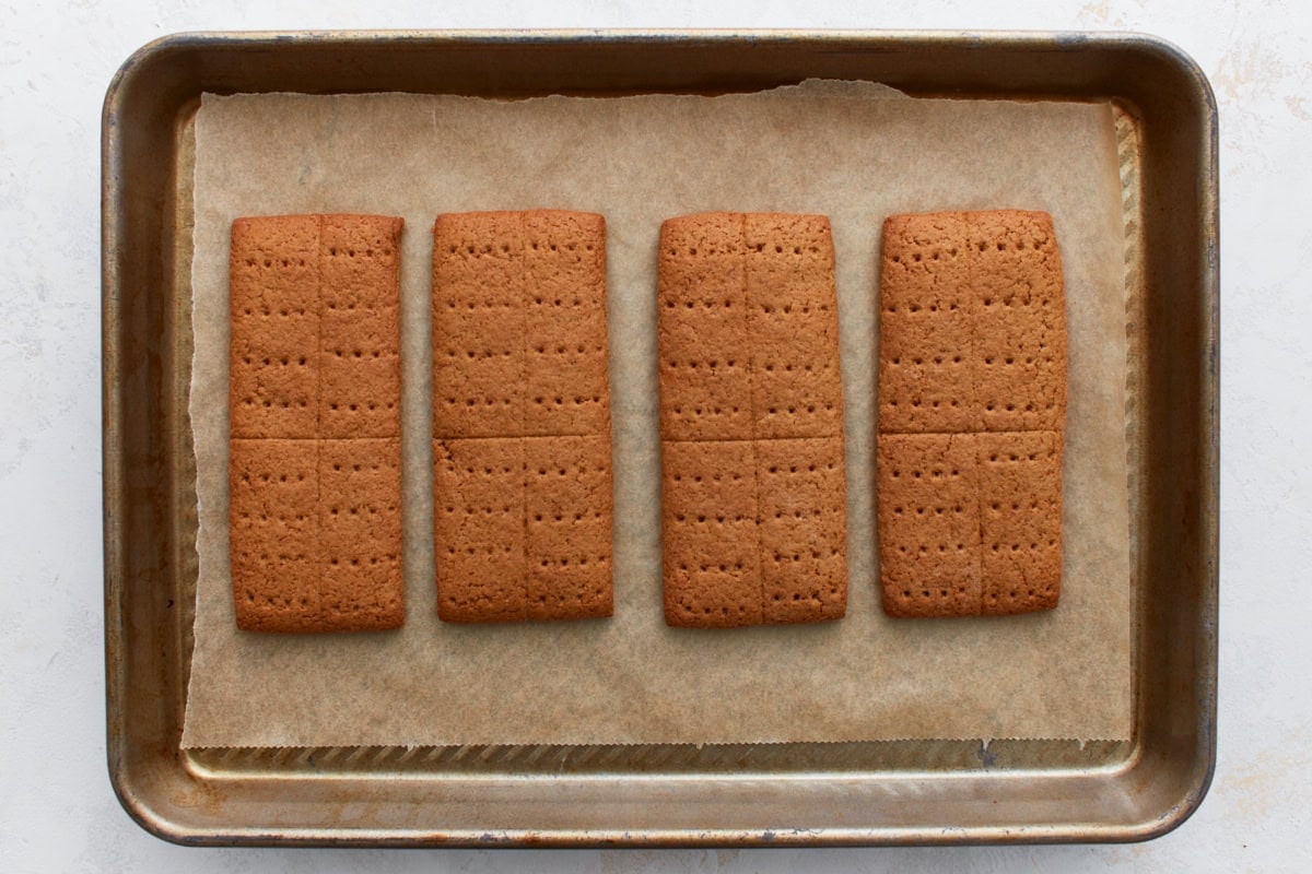 A baking sheet with freshly baked gluten-free graham crackers, golden brown and fully set, with their scored lines and fork pricks visible.