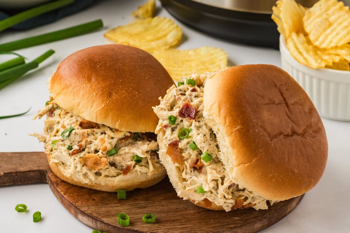 Two soft sandwich buns filled with creamy Instant Pot crack chicken, topped with chopped bacon and green onions, served on a wooden board with potato chips in the background.