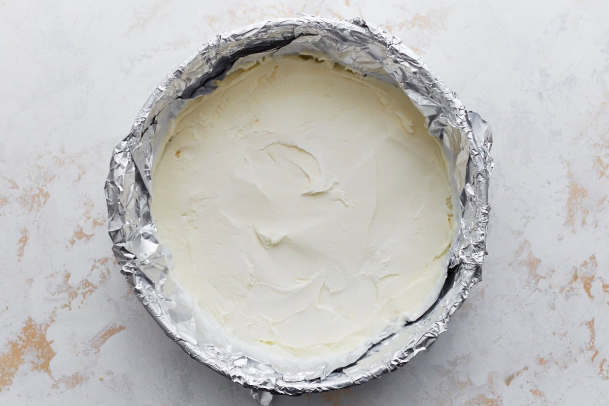 A round cake pan lined with foil and filled with smoothed vanilla gluten-free ice cream, ready to be frozen into a solid layer.