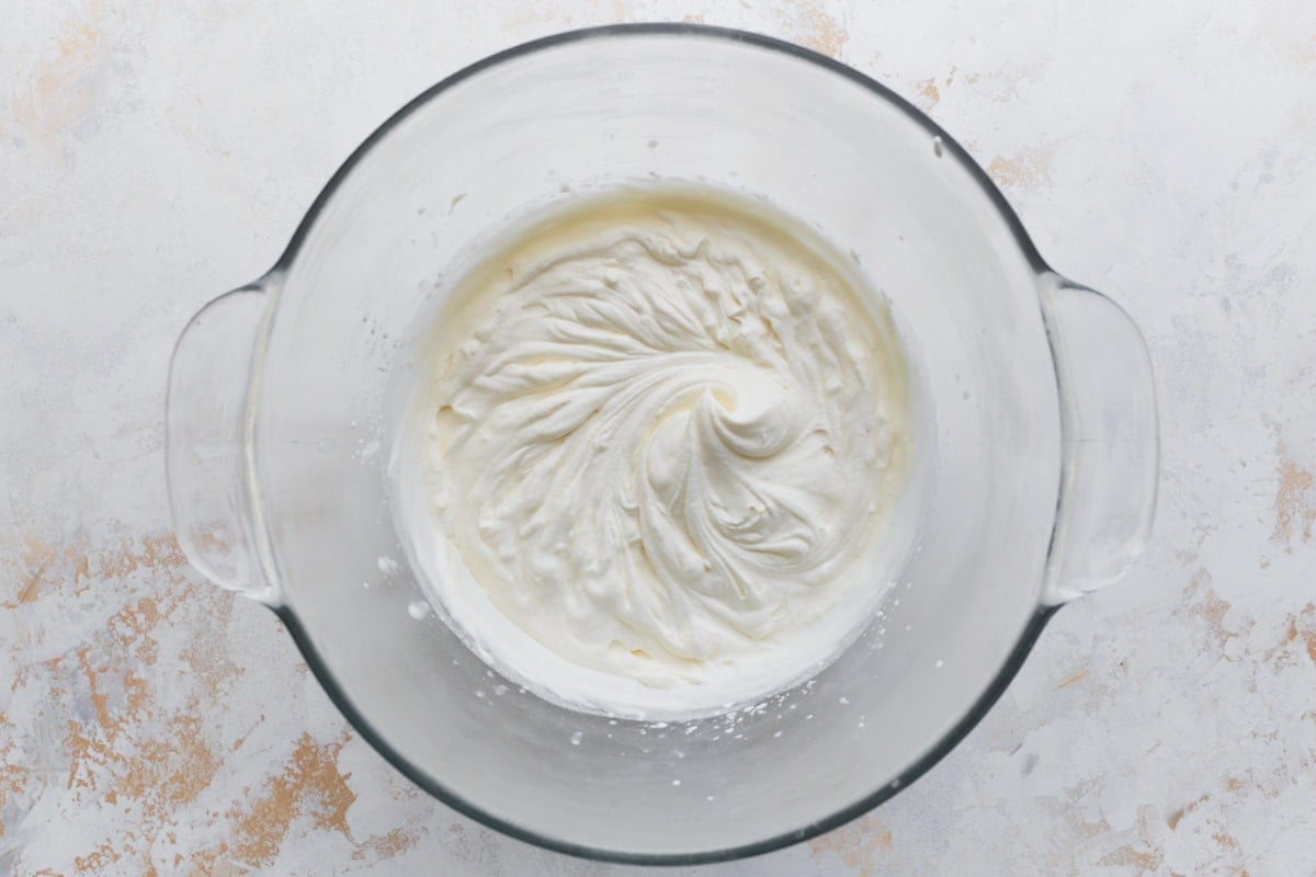 A glass mixing bowl with freshly whipped cream, showing soft, glossy peaks after mixing with vanilla, powdered sugar, and cornstarch.