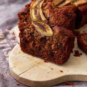 A close-up of a sliced loaf of banana oatmeal bread with dark chocolate chips and a caramelized banana topping, sitting on a wooden board.
