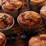 Close-up of golden-brown gluten-free blueberry muffins on a black wire cooling rack, surrounded by fresh blueberries.