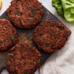 Four cooked gluten-free black bean burger patties on a rustic wooden board, surrounded by fresh lettuce and sandwich ingredients.