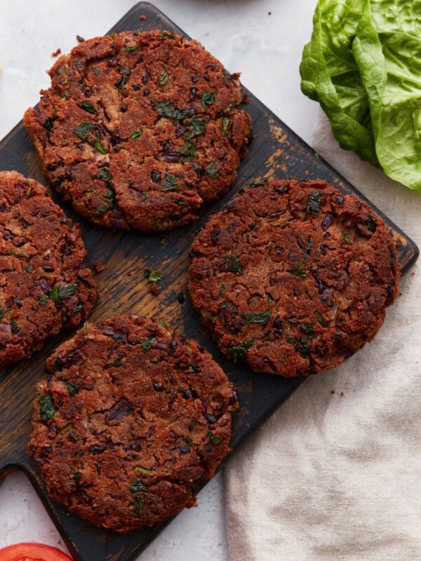 Four cooked gluten-free black bean burger patties on a rustic wooden board, surrounded by fresh lettuce and sandwich ingredients.
