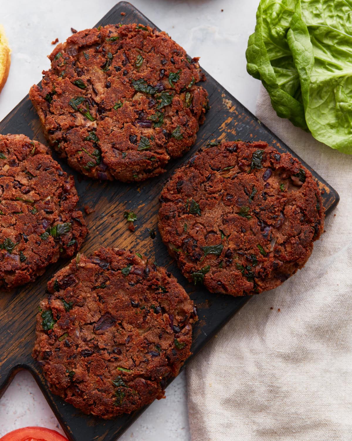 Four cooked gluten-free black bean burger patties on a rustic wooden board, surrounded by fresh lettuce and sandwich ingredients.