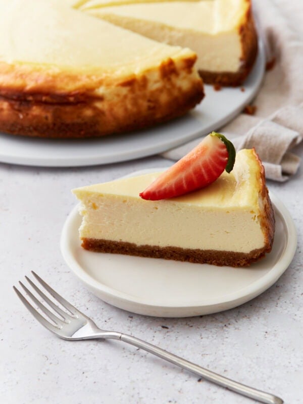 Slice of gluten-free cheesecake on a small white plate topped with a strawberry, with the rest of the cheesecake in the background.