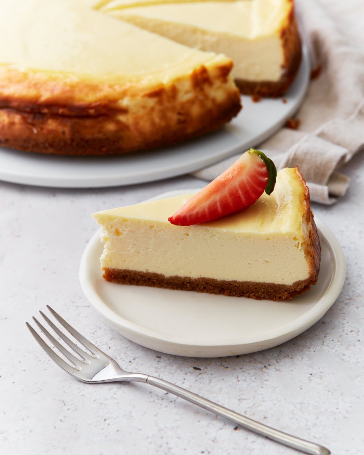Slice of gluten-free cheesecake on a small white plate topped with a strawberry, with the rest of the cheesecake in the background.

