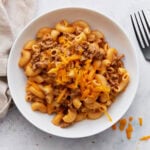 A white bowl filled with creamy gluten-free Hamburger Helper, topped with shredded cheddar cheese, sitting beside a fork on a light surface.