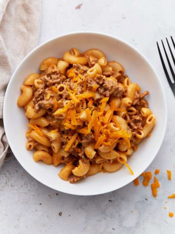 A white bowl filled with creamy gluten-free Hamburger Helper, topped with shredded cheddar cheese, sitting beside a fork on a light surface.