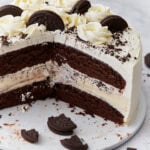 A layered gluten-free chocolate ice cream cake sliced to reveal chocolate cake layers, a vanilla ice cream center with cookie pieces, whipped cream frosting, and decorative chocolate cookies and rosettes on top.