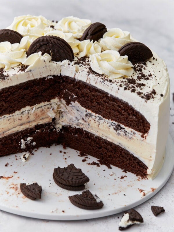 A layered gluten-free chocolate ice cream cake sliced to reveal chocolate cake layers, a vanilla ice cream center with cookie pieces, whipped cream frosting, and decorative chocolate cookies and rosettes on top.