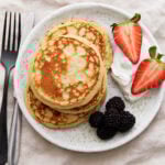 A stack of golden gluten-free protein pancakes on a white plate, served with blackberries, halved strawberries, and a dollop of Greek yogurt. A fork and knife sit beside the plate.