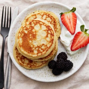 A stack of golden gluten-free protein pancakes on a white plate, served with blackberries, halved strawberries, and a dollop of Greek yogurt. A fork and knife sit beside the plate.