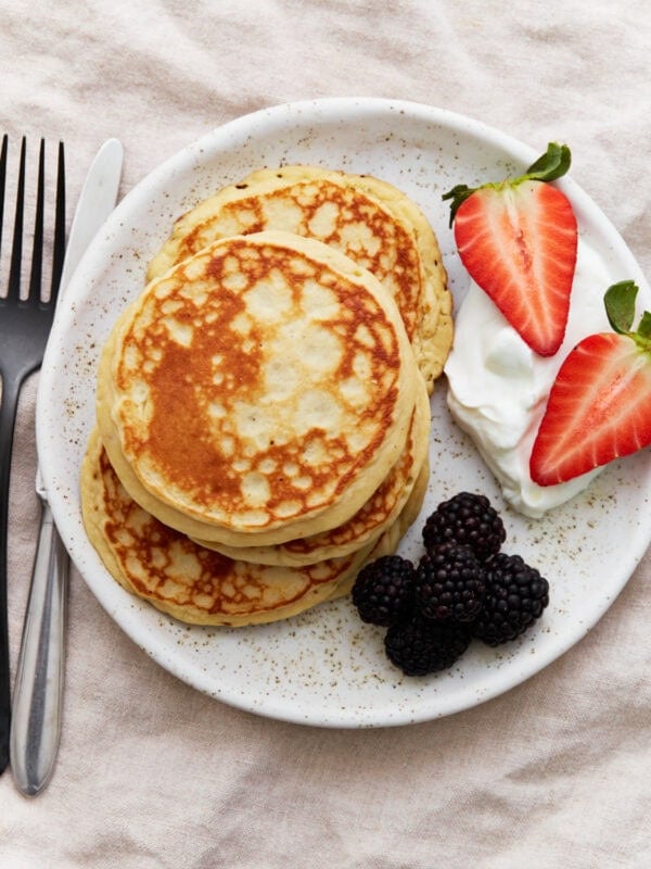 A stack of golden gluten-free protein pancakes on a white plate, served with blackberries, halved strawberries, and a dollop of Greek yogurt. A fork and knife sit beside the plate.