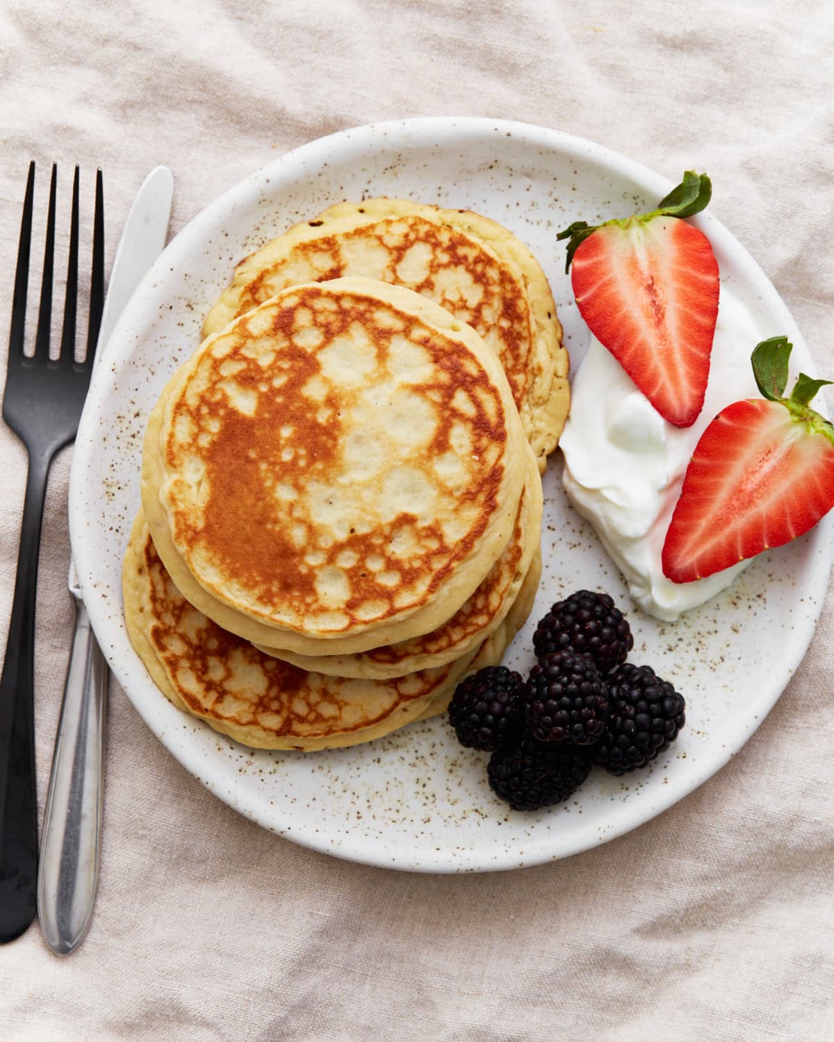 A stack of golden gluten-free protein pancakes on a white plate, served with blackberries, halved strawberries, and a dollop of Greek yogurt. A fork and knife sit beside the plate.