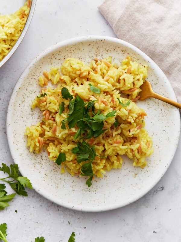 A serving of golden gluten-free rice pilaf garnished with chopped parsley on a speckled white plate with a gold fork.
