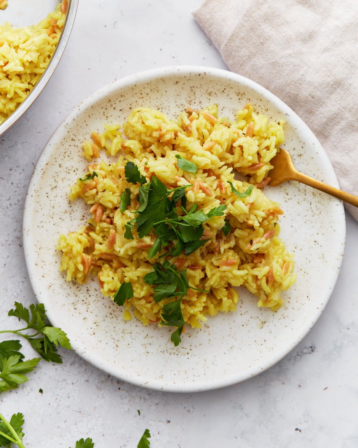 A serving of golden gluten-free rice pilaf garnished with chopped parsley on a speckled white plate with a gold fork.