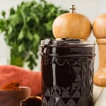 A glass jar filled with thick, dark homemade BBQ sauce, with sauce dripping slightly over the rim. Fresh parsley, garlic, and wooden pepper mills are in the background.