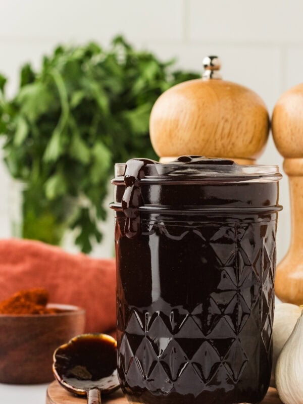 A glass jar filled with thick, dark homemade BBQ sauce, with sauce dripping slightly over the rim. Fresh parsley, garlic, and wooden pepper mills are in the background.