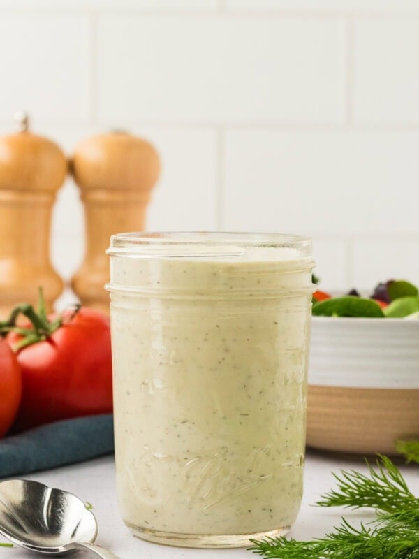 A glass mason jar filled with creamy dairy-free ranch dressing, set on a white countertop with tomatoes, fresh dill, and a spoon nearby.