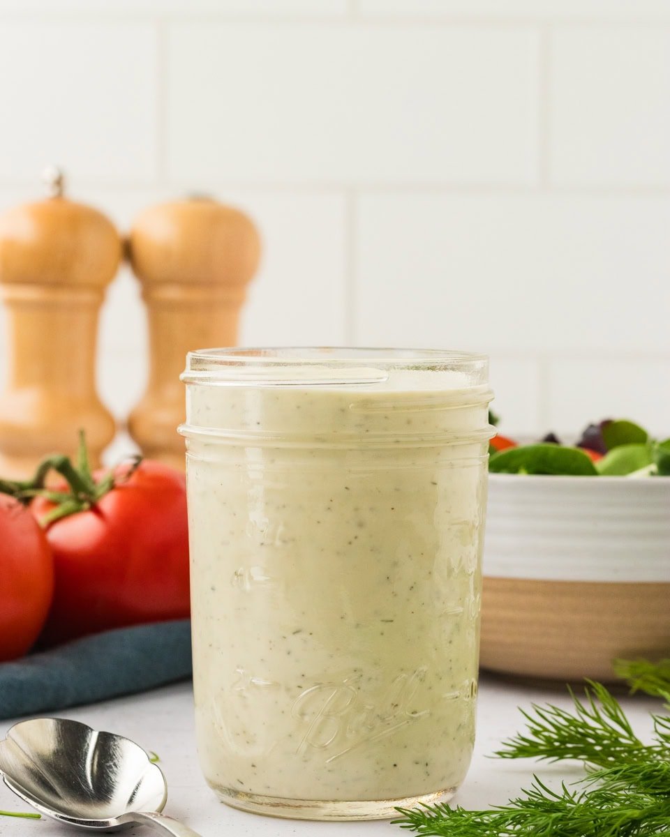 A glass mason jar filled with creamy dairy-free ranch dressing, set on a white countertop with tomatoes, fresh dill, and a spoon nearby.