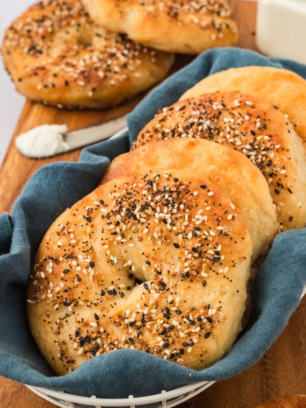 Golden brown gluten-free everything bagels arranged in a white basket lined with a blue cloth, served on a wooden board with a spread of cream cheese.