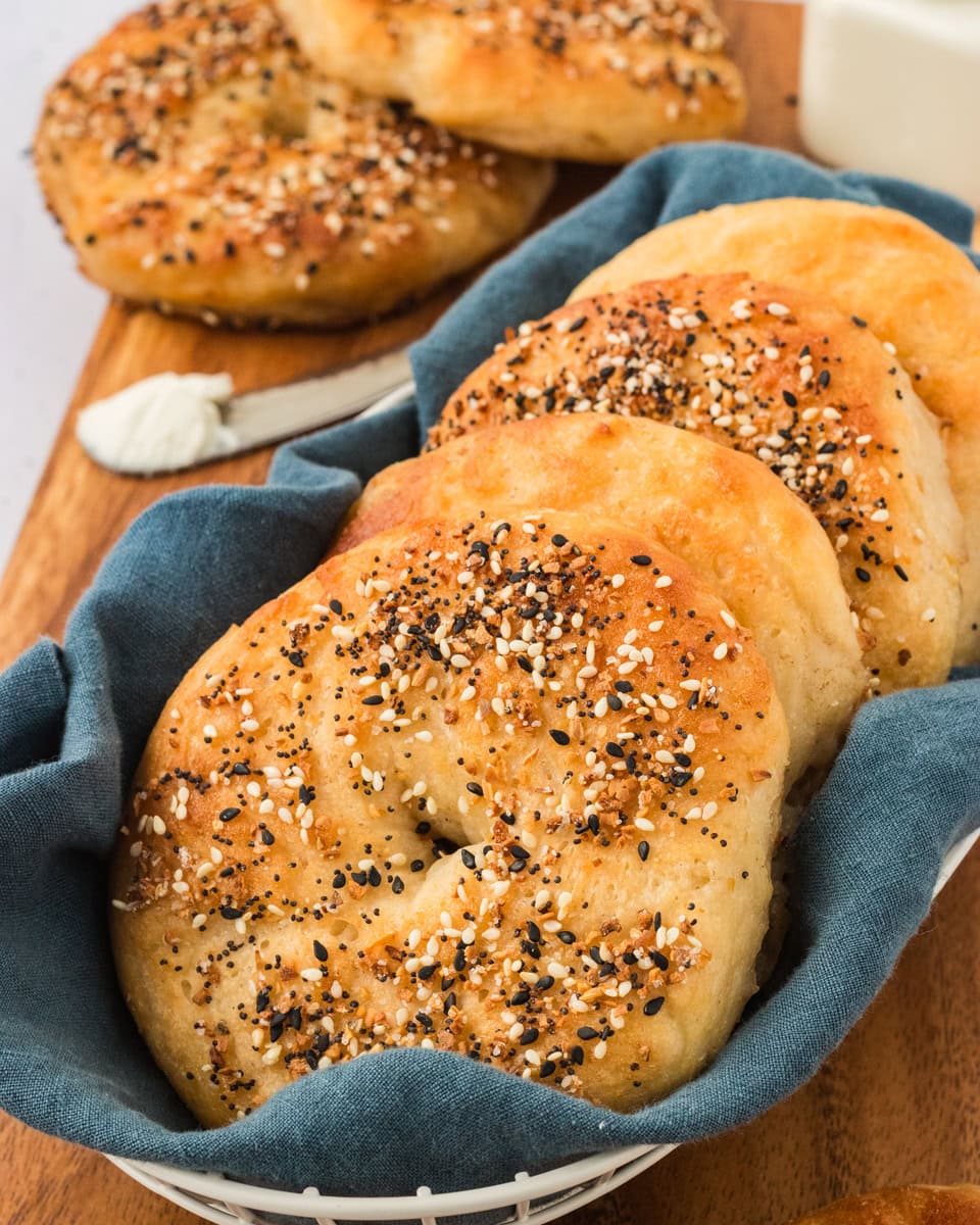 Golden brown gluten-free everything bagels arranged in a white basket lined with a blue cloth, served on a wooden board with a spread of cream cheese.