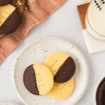 Two chocolate-dipped gluten-free shortbread cookies on a speckled plate next to a glass of milk with striped paper straws, perfect for serving.
