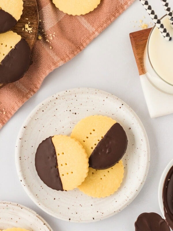 Two chocolate-dipped gluten-free shortbread cookies on a speckled plate next to a glass of milk with striped paper straws, perfect for serving.