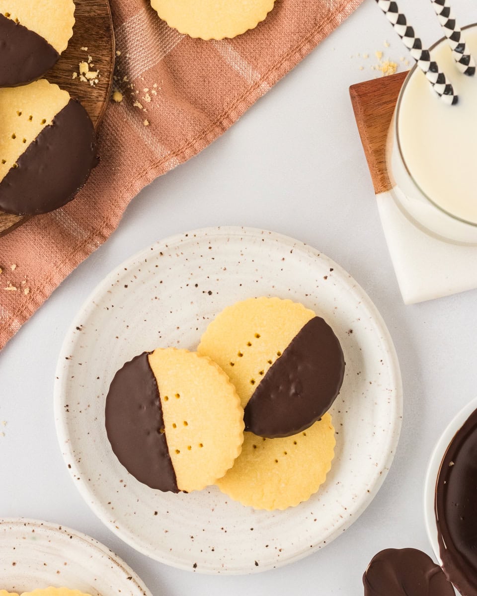 Two chocolate-dipped gluten-free shortbread cookies on a speckled plate next to a glass of milk with striped paper straws, perfect for serving.
