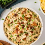 Bowl of creamy crack chicken topped with chopped green onions and crispy bacon, surrounded by fresh toppings and chips.