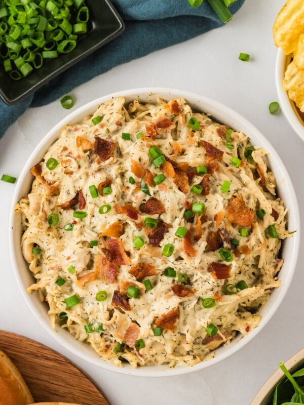 Bowl of creamy crack chicken topped with chopped green onions and crispy bacon, surrounded by fresh toppings and chips.