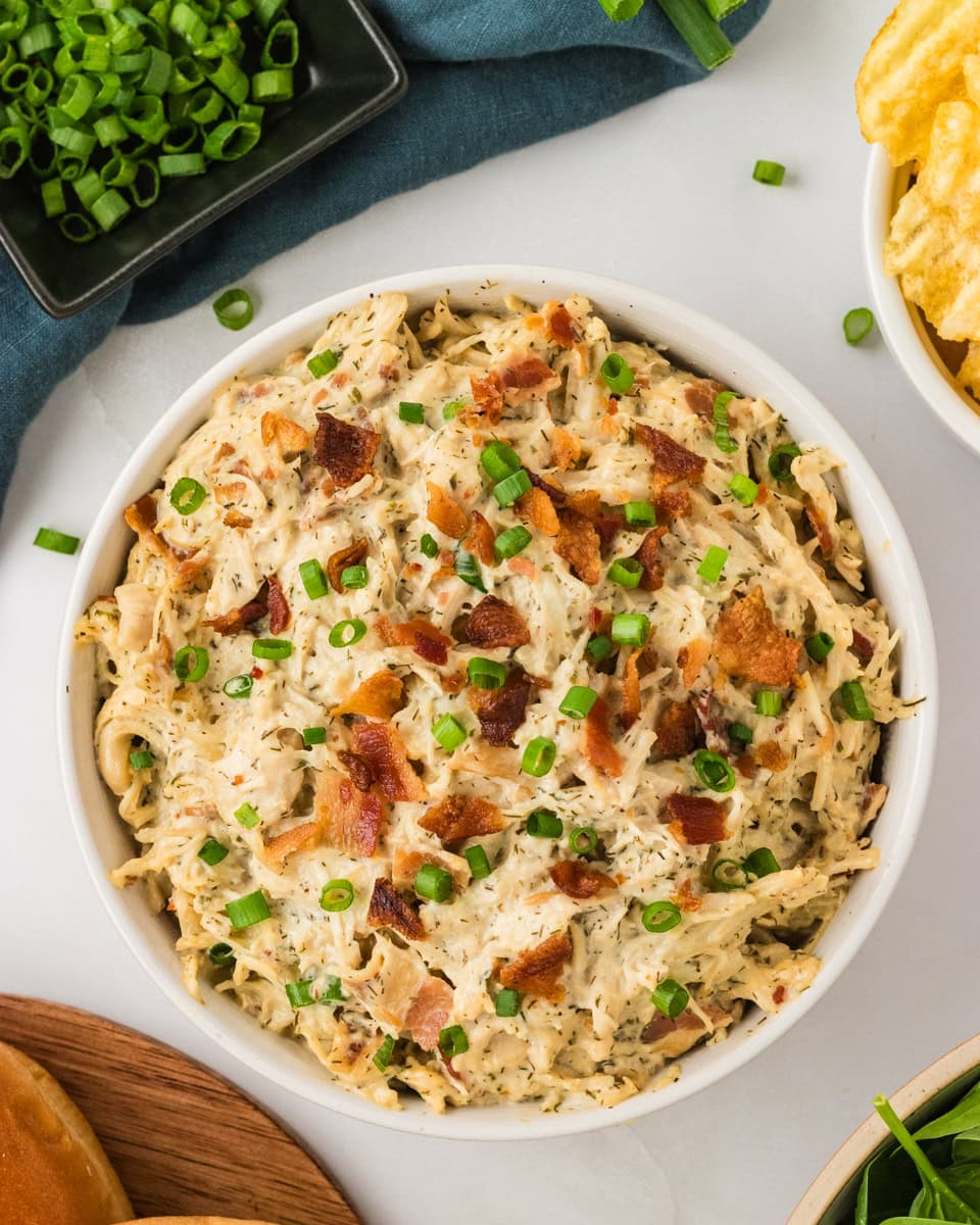 Bowl of creamy crack chicken topped with chopped green onions and crispy bacon, surrounded by fresh toppings and chips.