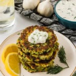 A stack of crispy zucchini fritters topped with a dollop of creamy yogurt sauce and garnished with fresh dill, served with lemon wedges on a white plate.