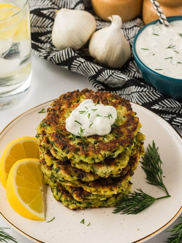 A stack of crispy zucchini fritters topped with a dollop of creamy yogurt sauce and garnished with fresh dill, served with lemon wedges on a white plate.