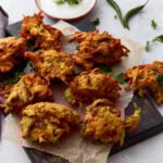 Golden-brown pakoras arranged on parchment paper over a dark wooden board, served with a bowl of creamy white dipping sauce and fresh herbs.