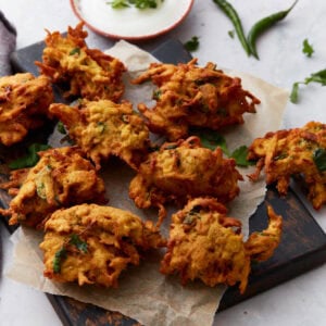 Golden-brown pakoras arranged on parchment paper over a dark wooden board, served with a bowl of creamy white dipping sauce and fresh herbs.