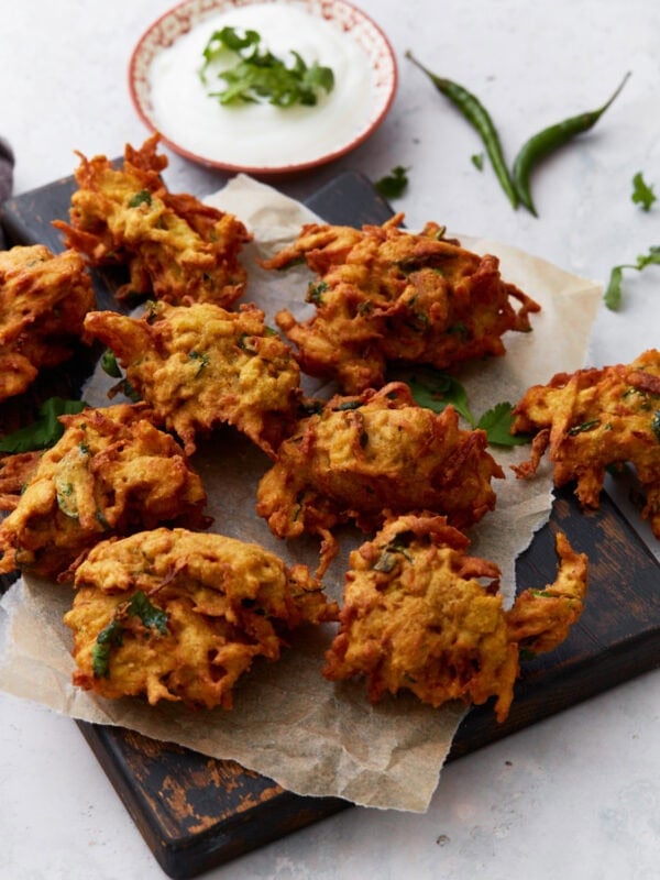 Golden-brown pakoras arranged on parchment paper over a dark wooden board, served with a bowl of creamy white dipping sauce and fresh herbs.