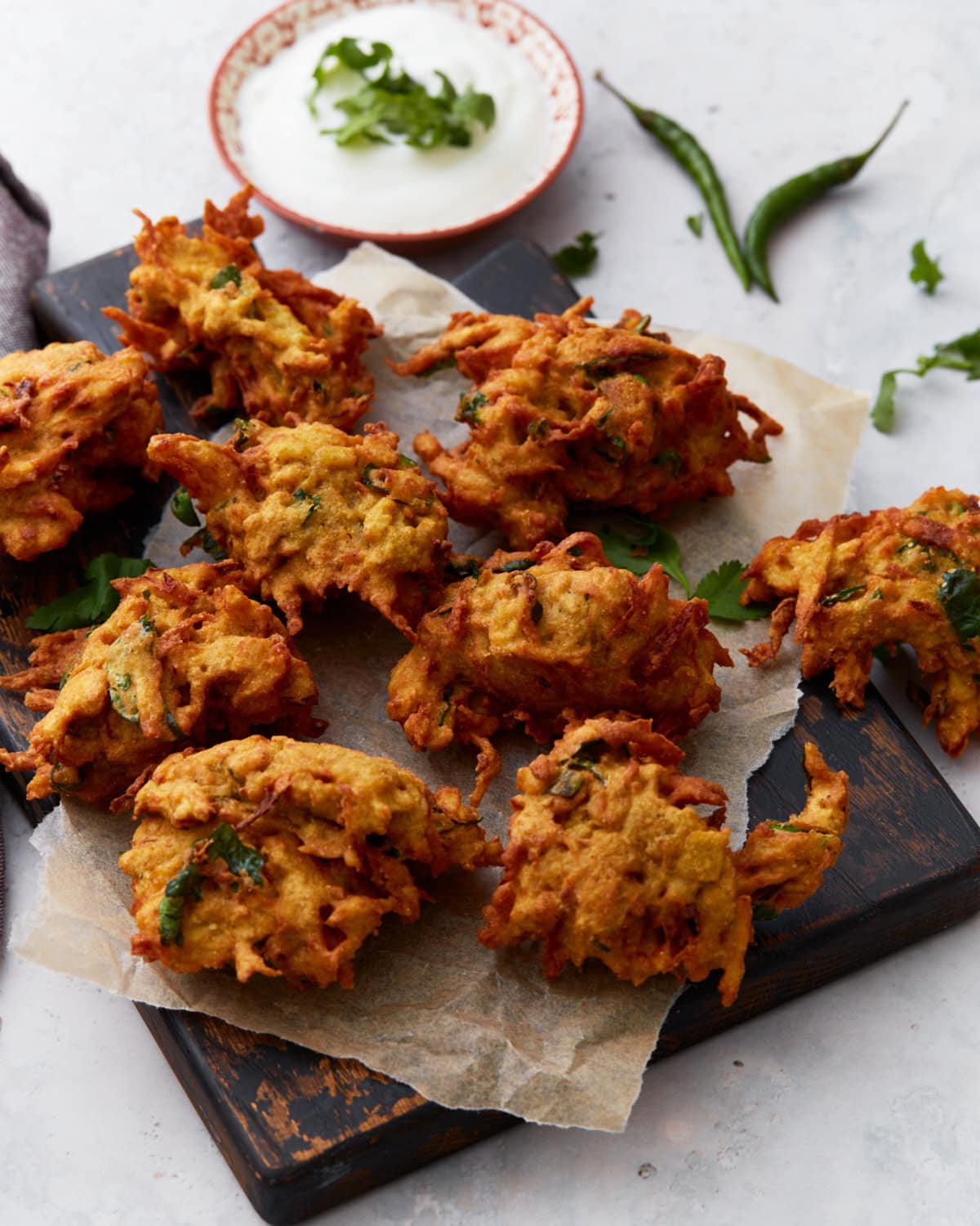 Golden-brown pakoras arranged on parchment paper over a dark wooden board, served with a bowl of creamy white dipping sauce and fresh herbs.