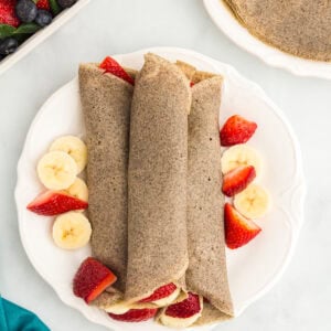 Three rolled buckwheat crepes on a white plate, surrounded by fresh banana slices and halved strawberries.
