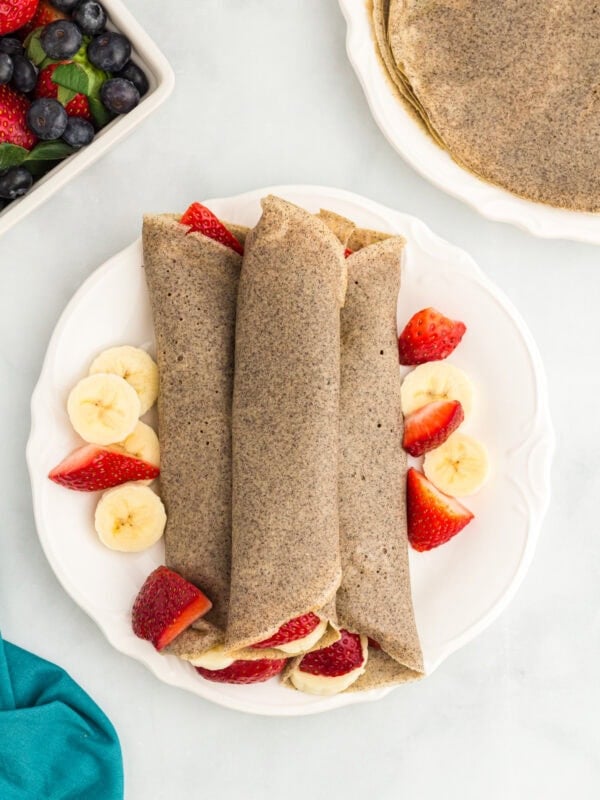 Three rolled buckwheat crepes on a white plate, surrounded by fresh banana slices and halved strawberries.