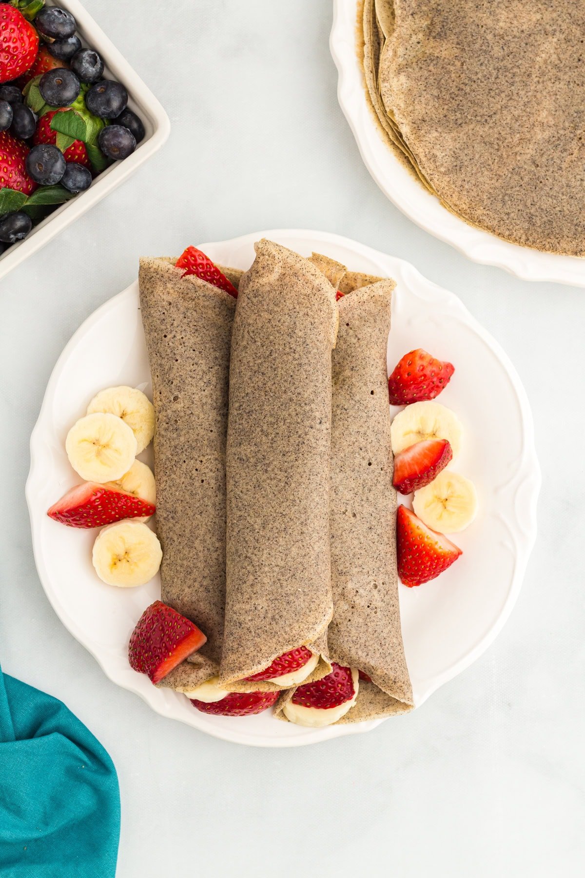Three rolled buckwheat crepes on a white plate, surrounded by fresh banana slices and halved strawberries.