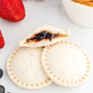 Two gluten-free uncrustables stacked with one cut open to show the peanut butter and blueberry jam filling, surrounded by fresh strawberries, blueberries, and a small bowl of peanut butter.