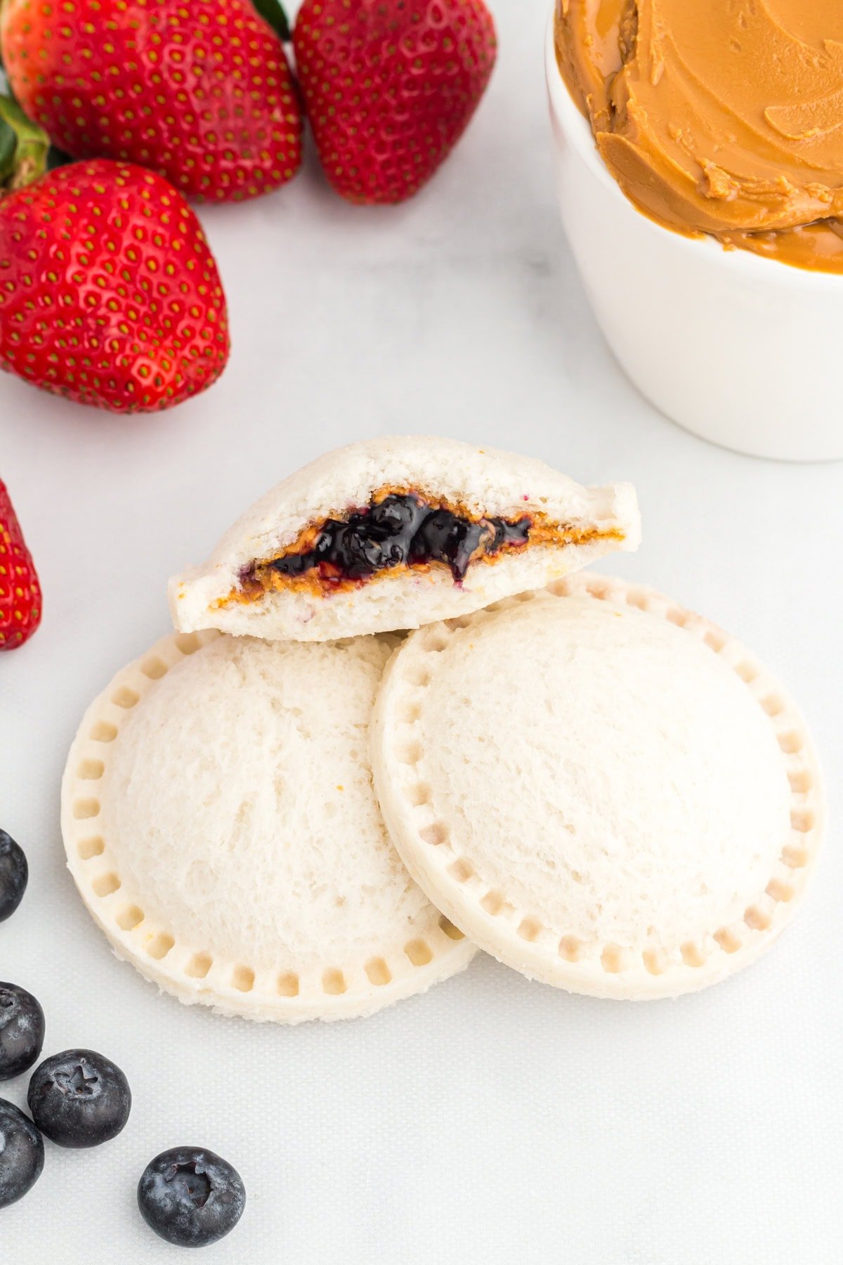 Two gluten-free uncrustables stacked with one cut open to show the peanut butter and blueberry jam filling, surrounded by fresh strawberries, blueberries, and a small bowl of peanut butter.