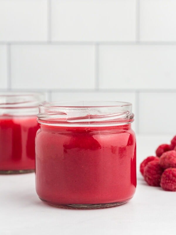 Close-up of a glass jar filled with vibrant red raspberry coulis, with fresh raspberries beside it on a white surface and a white tiled background.