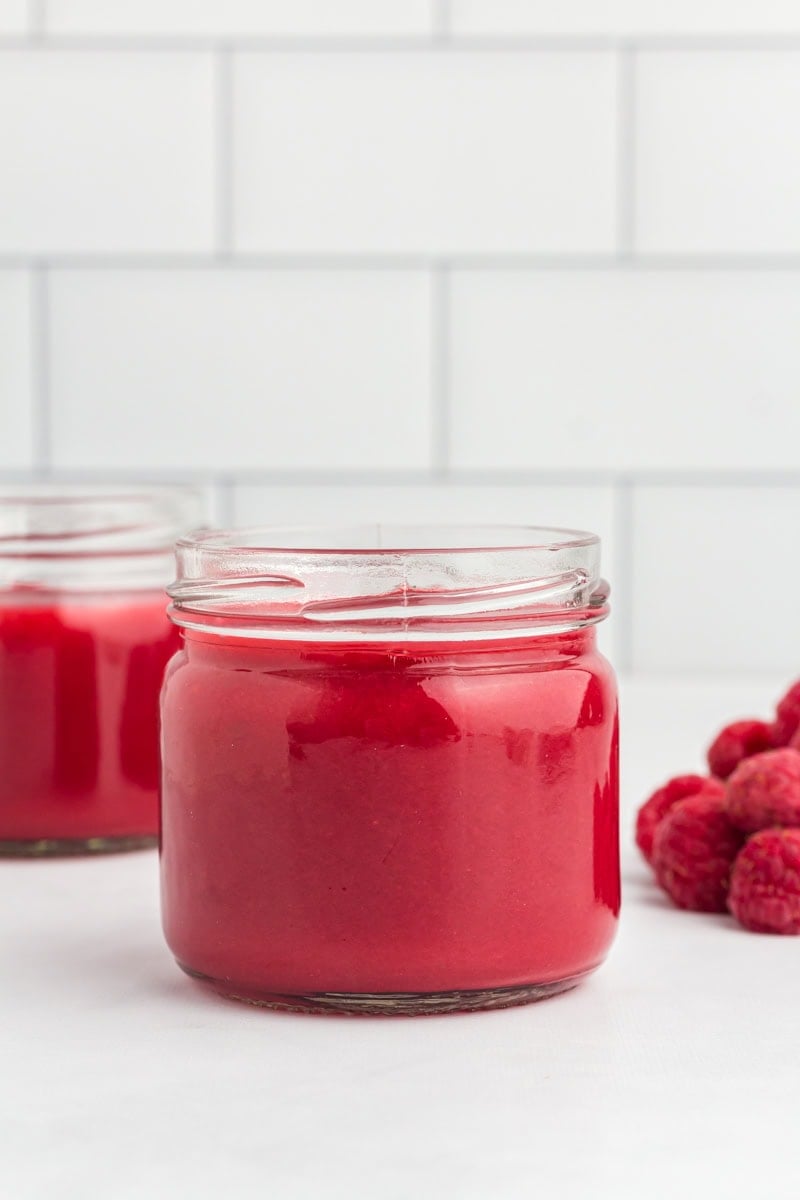 Close-up of a glass jar filled with vibrant red raspberry coulis, with fresh raspberries beside it on a white surface and a white tiled background.