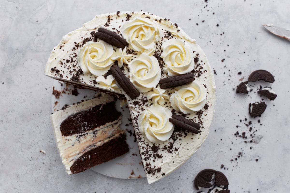 A sliced gluten-free ice cream cake showing the interior layers of chocolate cake, ice cream with cookie pieces, and whipped cream frosting, with cookie crumbs scattered on the surface.