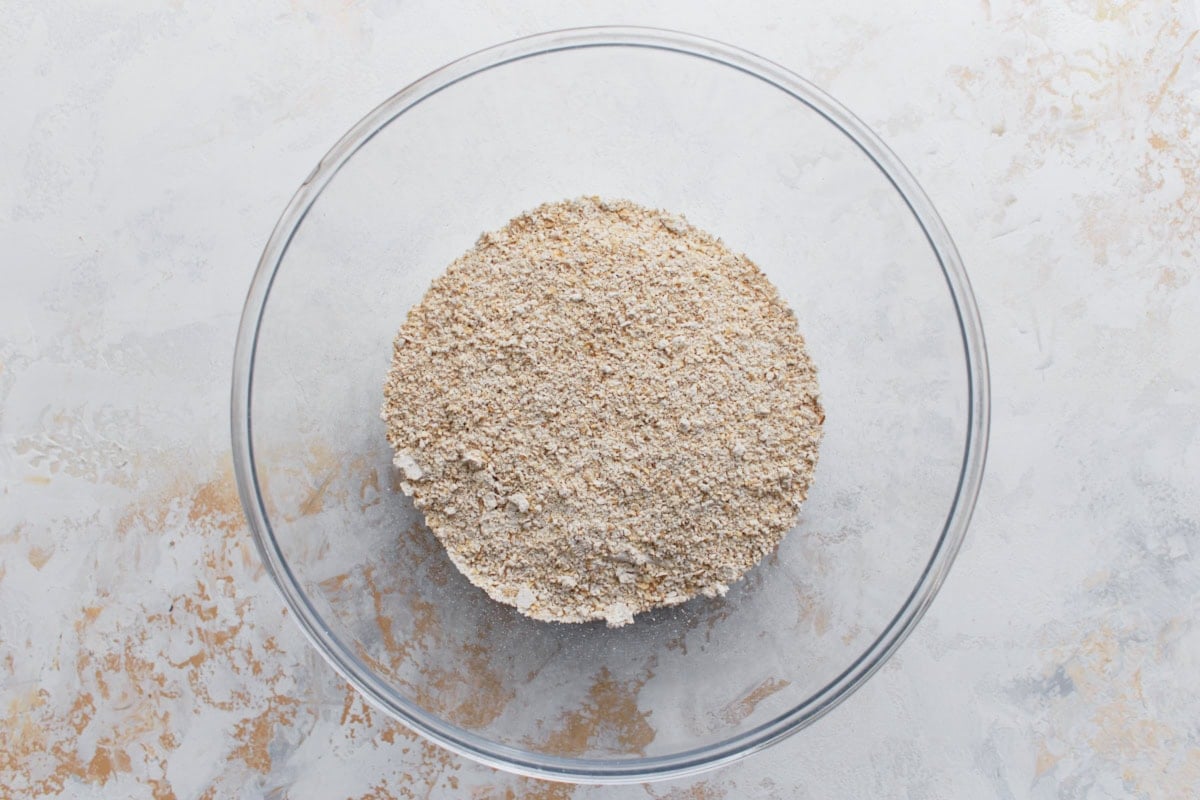 A glass bowl filled with freshly blended oat flour, made from rolled oats, on a light textured surface.